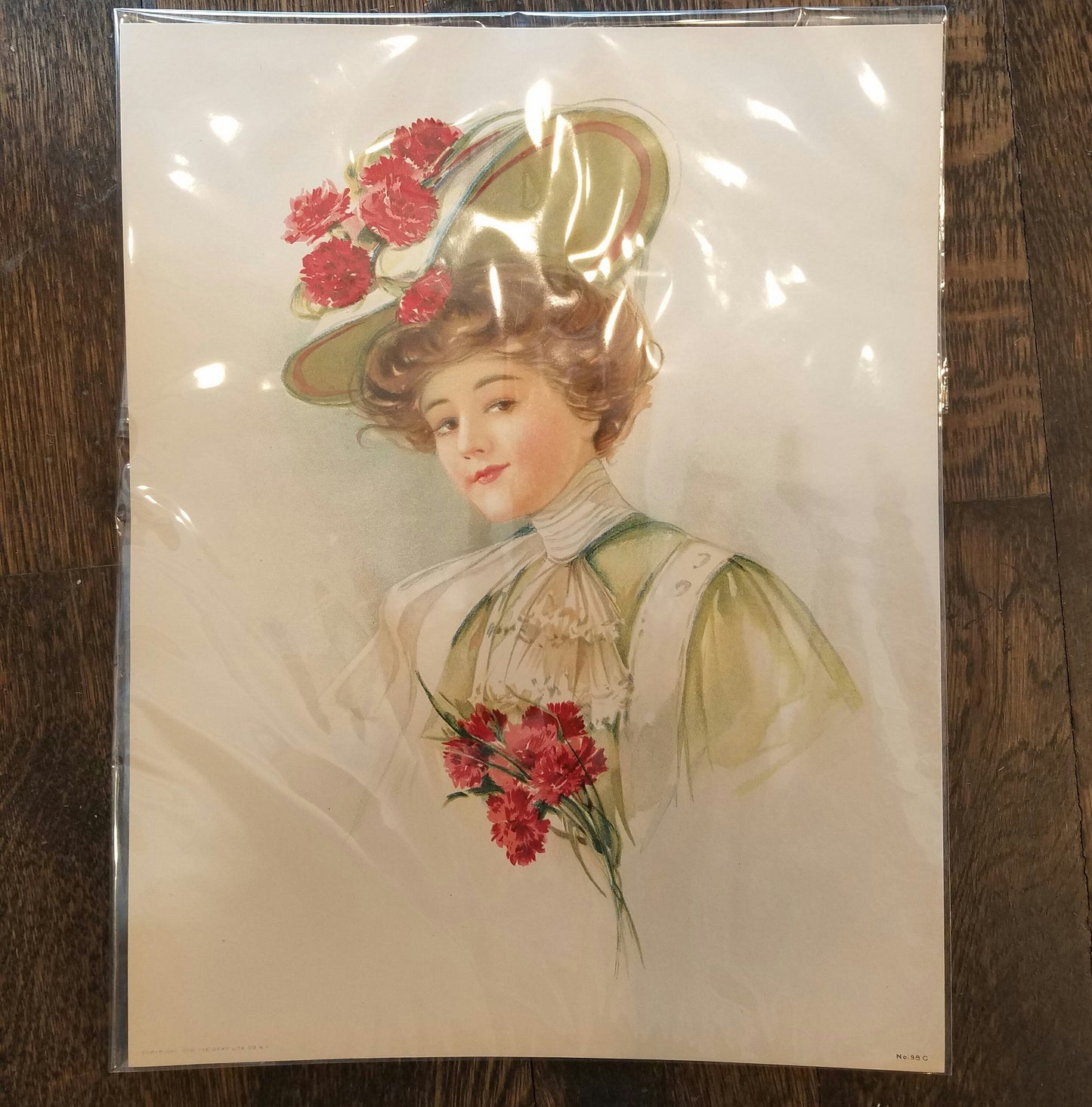 Victorian illustration of a brunette woman with brown eyes. Her hair is tied upward in a mint green, wide brimmed fancy hat with carnations on top of it. She has a high lace collar and ruffled lace bib of cream on her mint green dress. She is holding carnations.