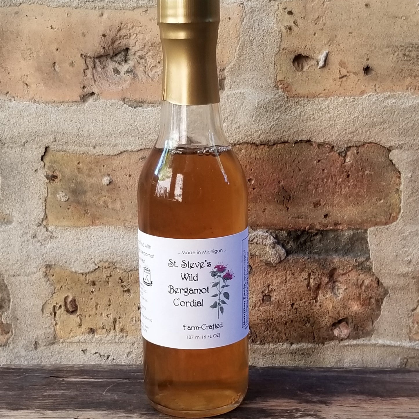 A clear glass, tall, thin bottle with an amber liquid. Bottle sits on wood and bricks are in background.