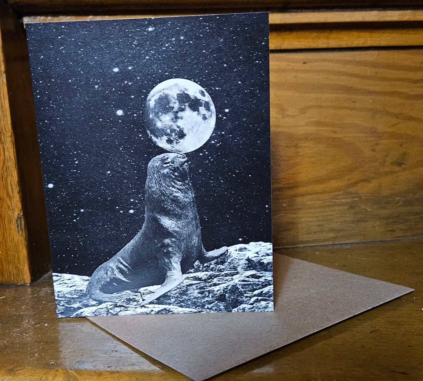 vintage collage grainy black and white image of a seal balancing the moon on their nose. They are standing on a rocky surface and the stars are twinkling behind them. The card is blank inside and the envelope is craft brown.