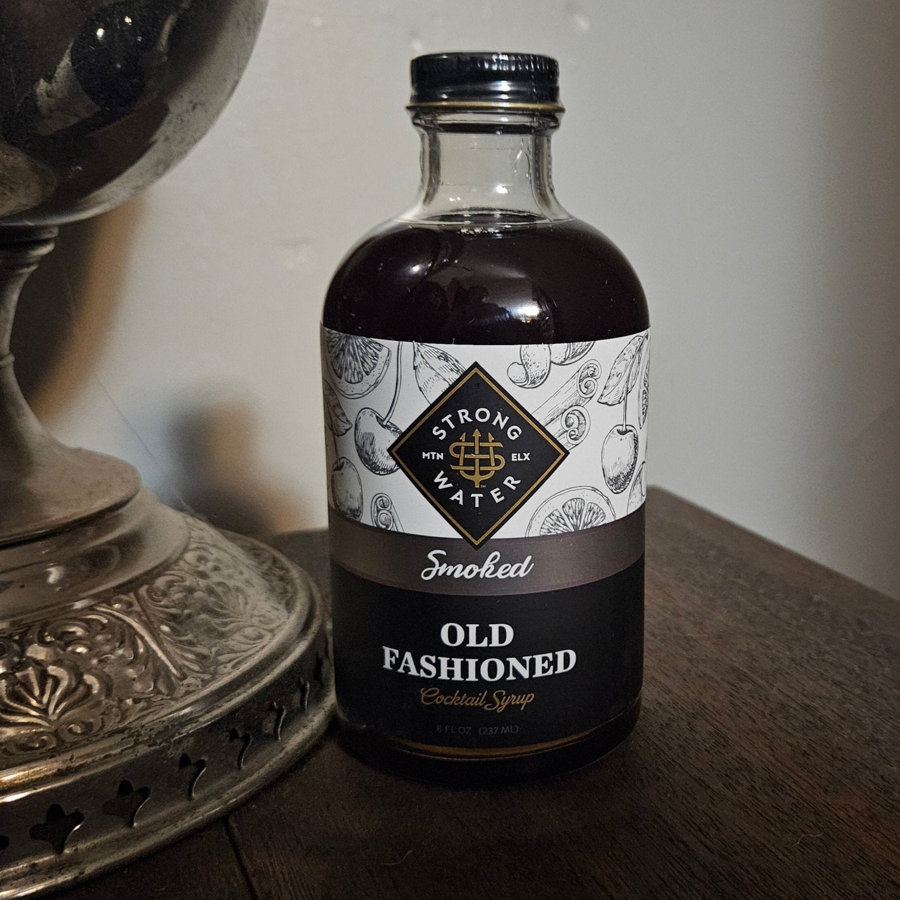 a round glass clear bottle with a dark brown liquid. label is white, copper, black, and metallic gold. bottle sits on walnut wood with antique silver hurricane lamp next to it. wall in back is pale grey.