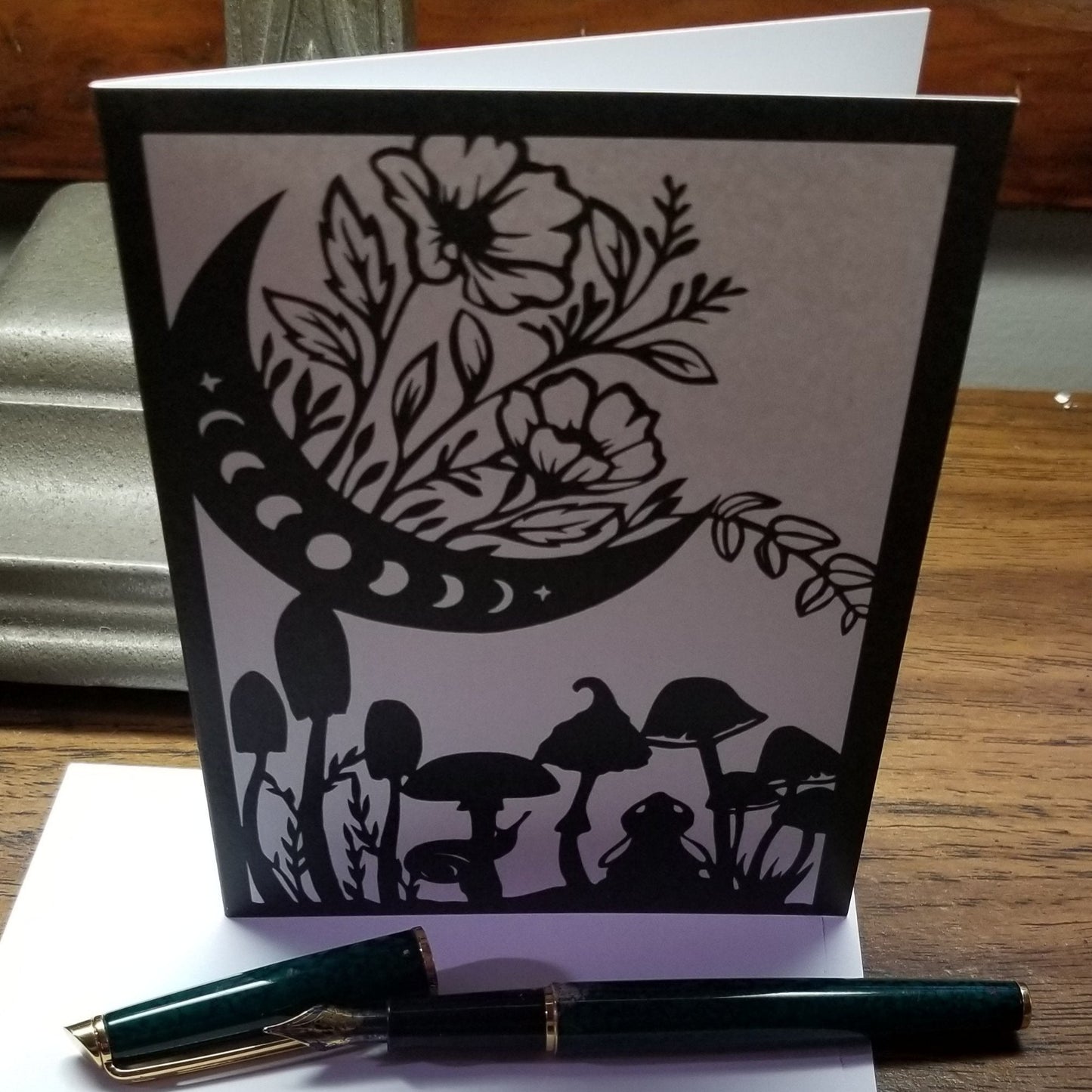 black and white card with outlines of a crescent moon with moonphase design, a bunny looking up at the moon, mushrooms, and flowers.