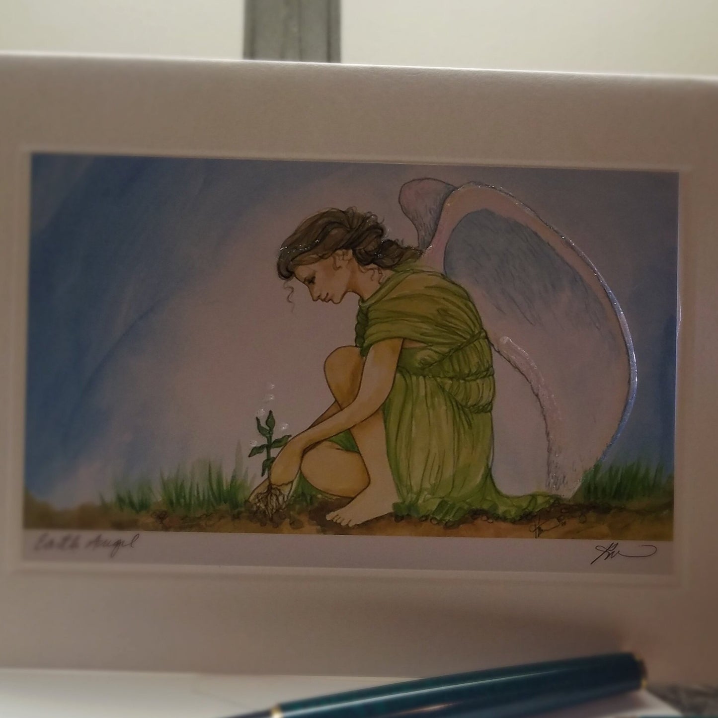 White card, oriented landscape, with print of kneeling, white winged fairy. She has long brown hair, tied back, and is wearing a gauzy green dress. She is planting  tree and sits in dirt with sprouting green. Background is blue sky. Card is hand detailed in iridescent glitter around angel wing and is hand signed by artist.