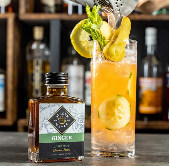 A short, rectangular clear glass bottle. Liquid is amber colored. Label is white, black, green, and metallic gold. Bottle sits on slate slab with bottles on shelf in background. Next to bottle is a highball cocktail which is orange colored with cucumbers and citrus floating in it.