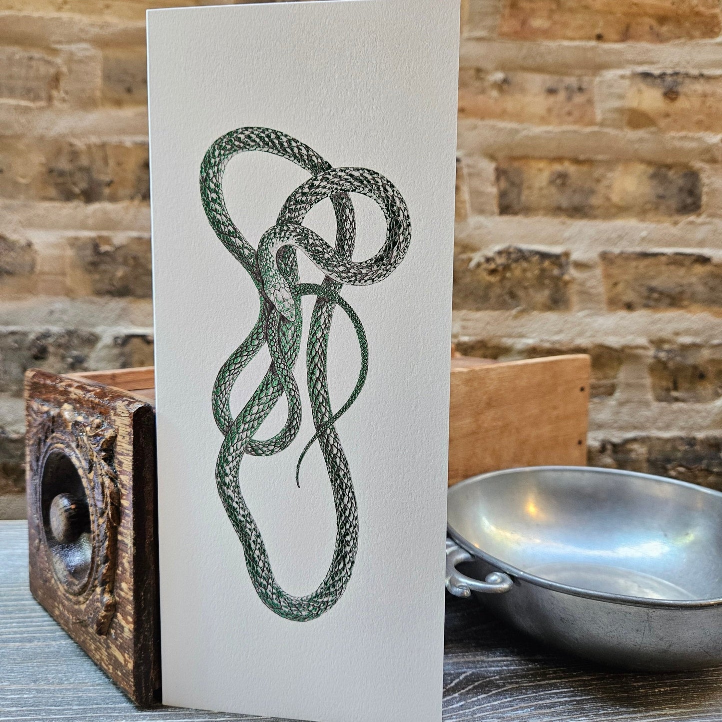 tall, white, rectangular card with foil metallic green illustration of green mamba snake coiled and twisted. background is old brick and props are antique wooden drawer and silver bowl. 