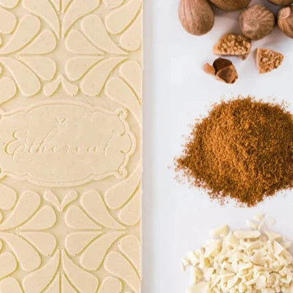 A white chocolate bar sits beside whole nutmeg, nutmeg powder, and shaved white chocolate