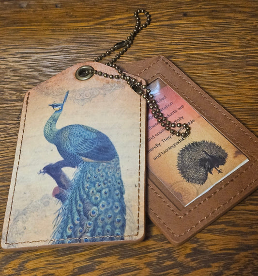 cognac brown luggage tag with brushed brass metal ball chain. Tag has vintage image of blue and green peacock with tail feathers downward and vertical plume on the top of their head. Peacock is sitting on a tree branch.