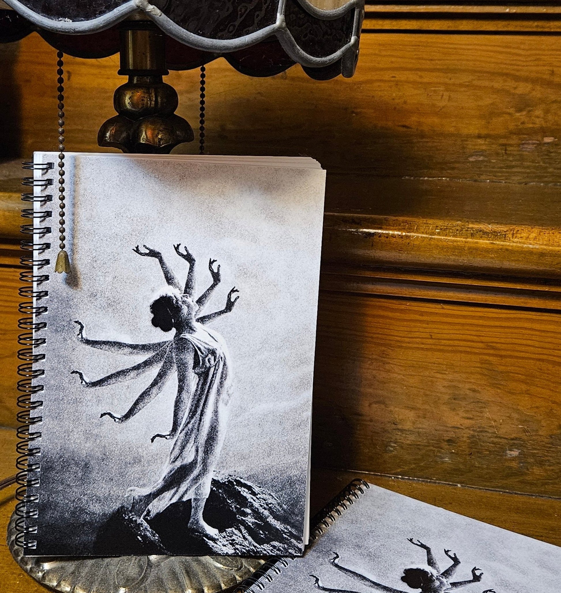 Spiral notebook with image on front and back cover. Black and white, vintage collage image features a goddess-like woman of the 1920s with 8 arms, standing on a mountain top. 