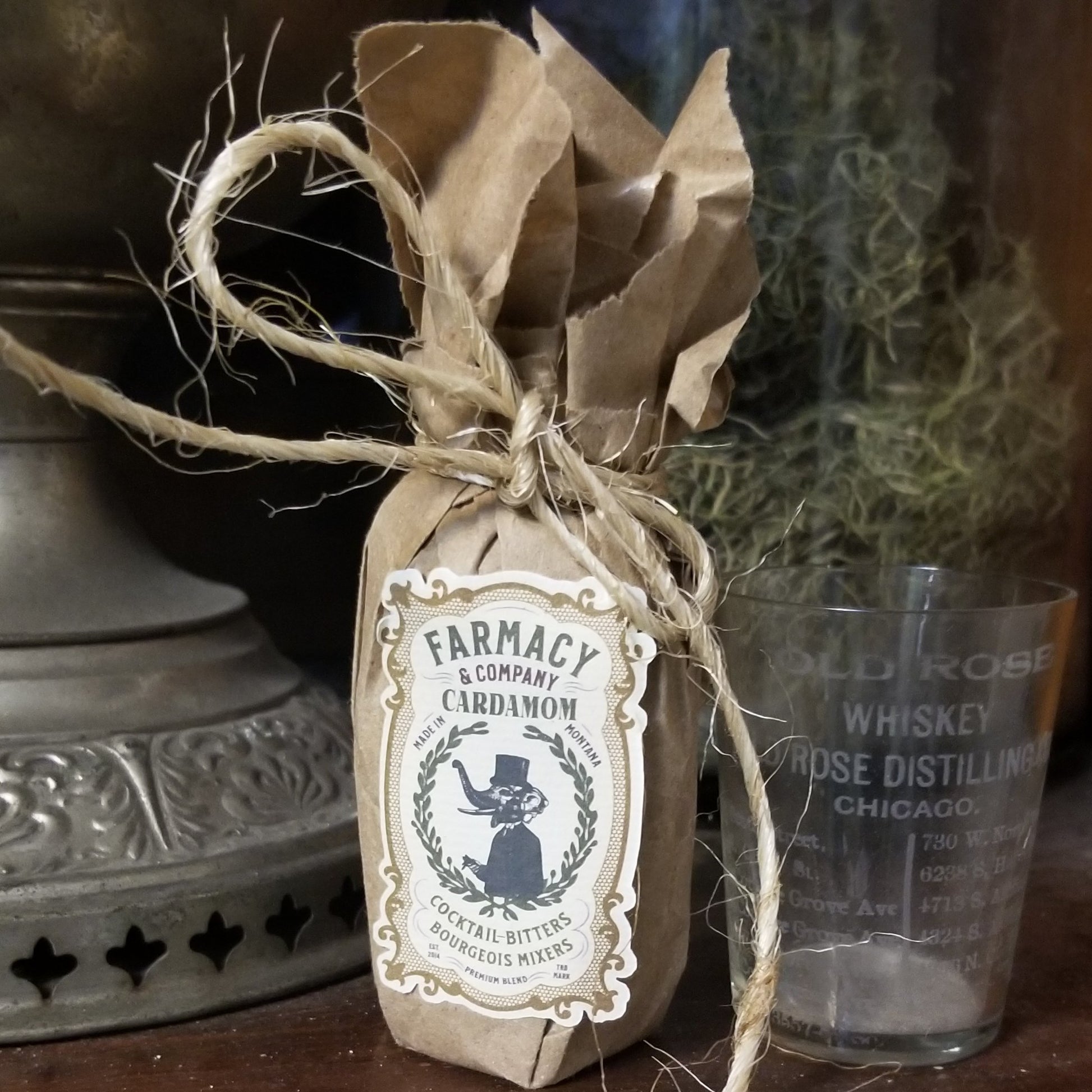 a small paper bag wrapped bottle, tied shut with a bow of jute. Label looks vintage and is an illustration of an elephant in suit and top hat. Background is an old hurricane lamp and an old rose distillery shot glass.