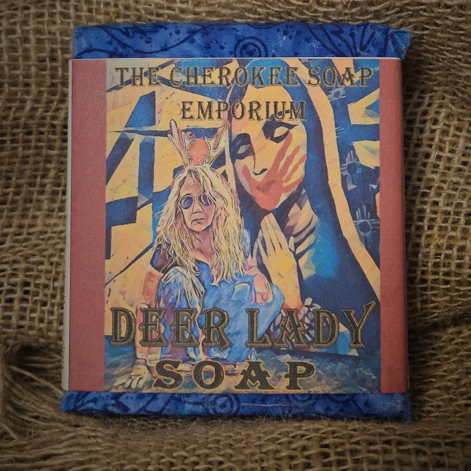 Bar of soap, wrapped in blue patterned Cherokee tear cloth with paper label featuring native artwork of a woman wearing sunglasses, crouching down. In the background, a Virgin Mary-like image prays with eyes closed and has a red hand paint print across her mouth and face. This is the symbol for Missing and Murdered Indigenous Women. 