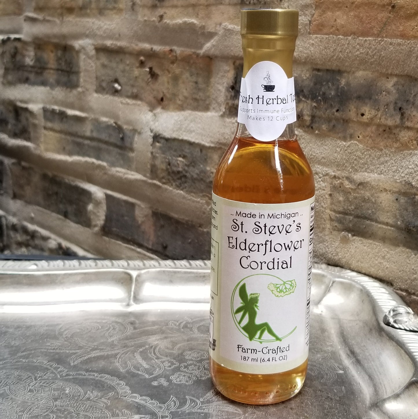 A clear, glass, tall, thin bottle with an amber colored liquid inside. Bottle sits on sterling silver serving tray and background is old bricks.