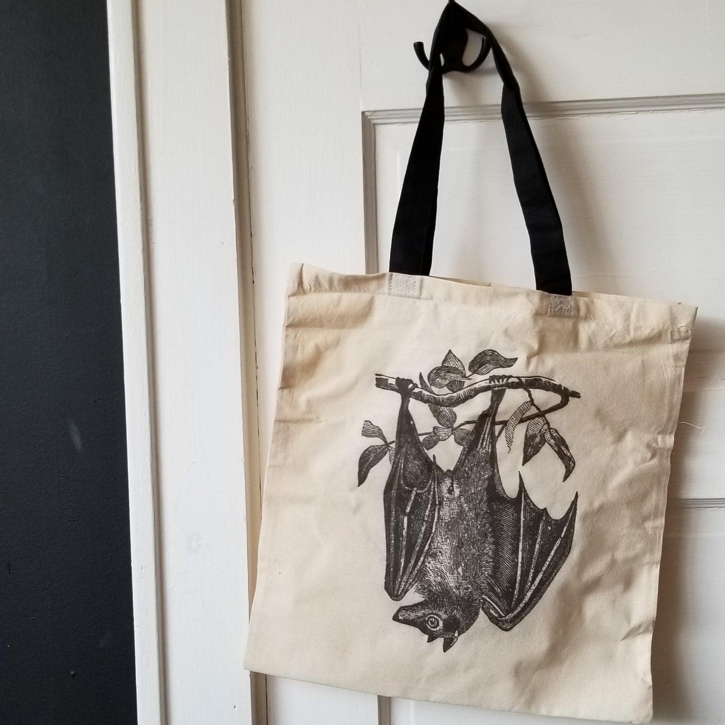 White door with black iron hook holding a cream, square, canvas totebag with a black handle. On the tote is a black illustration of a fruitbat hanging upside down from a branch. 