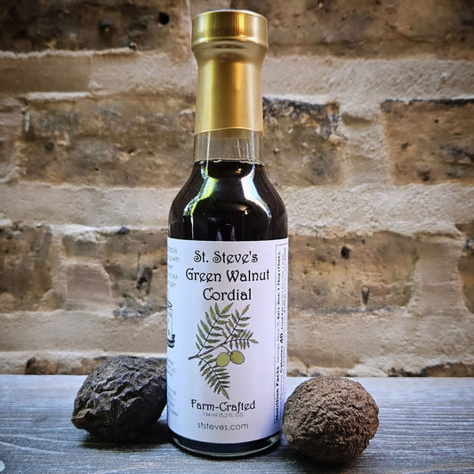 tall, thin, clear gloss bottle with a dark green liquid. bottle is wrapped in a dull gold seal at top. label is white with illustration of green walnuts on a walnut tree leaf. background image is brick and bottle sits on wood with two green walnuts beside it.