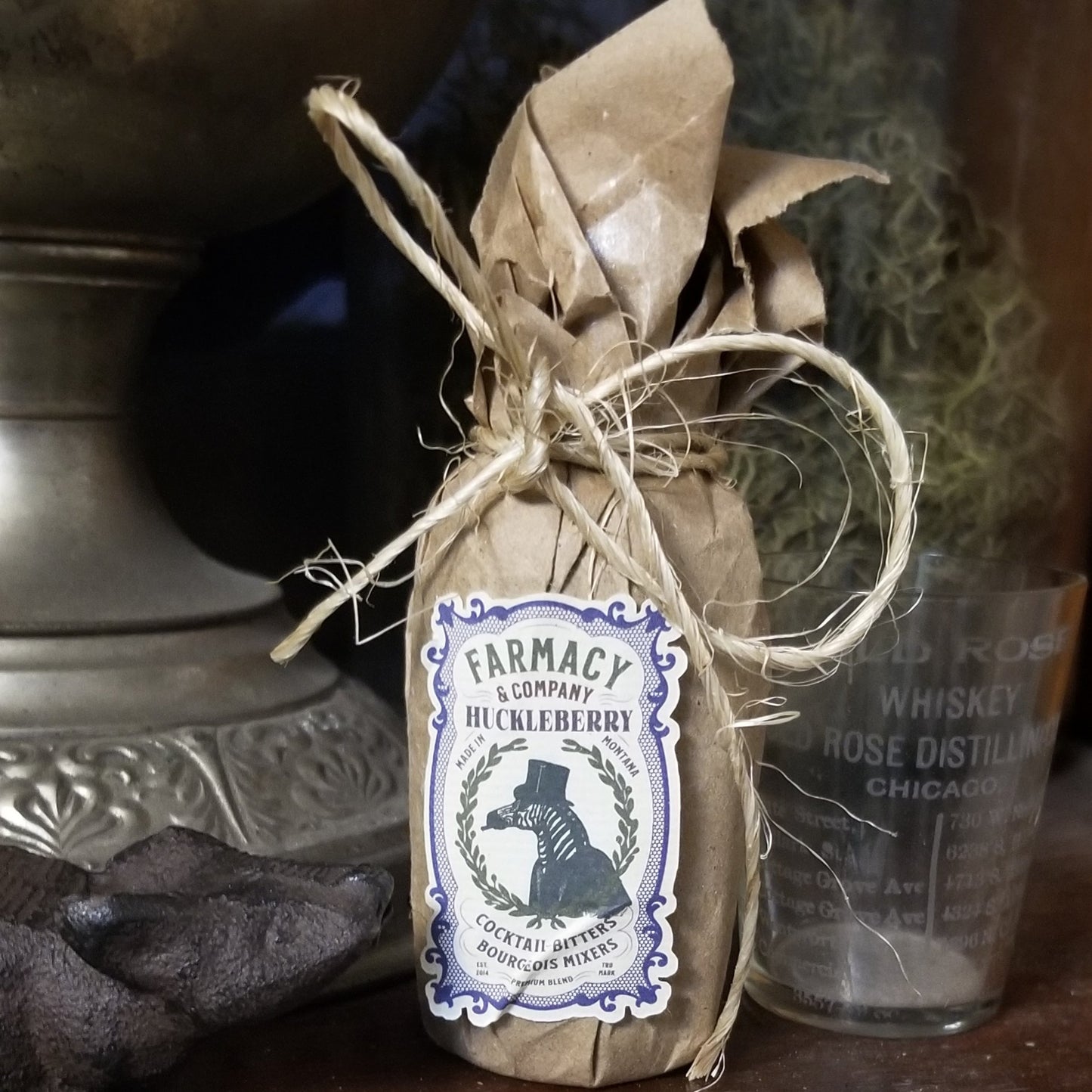 small bottle wrapped in craft paper and tied with a bow of jute. label is purple and black and cream and features a vintage-looking illustration of a zebra in a suit and top hat. Background is an old hurricane lamp and old rose distillery shot glass.