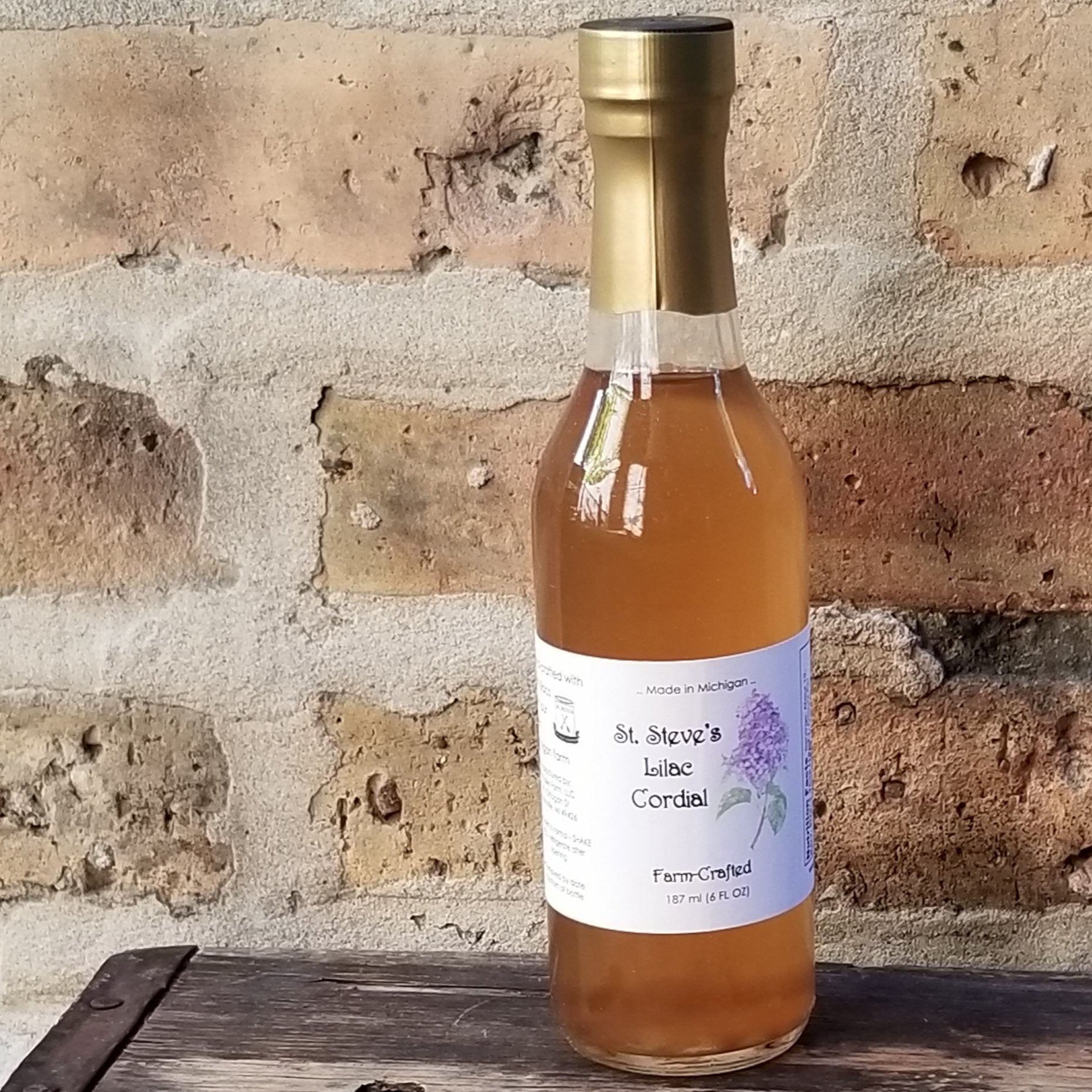 A tall, narrow, clear glass bottle with an amber liquid inside. Bottle has a dull gold wrapper at the top and a white label at the center with brand illustration. Background image is brick and wood.