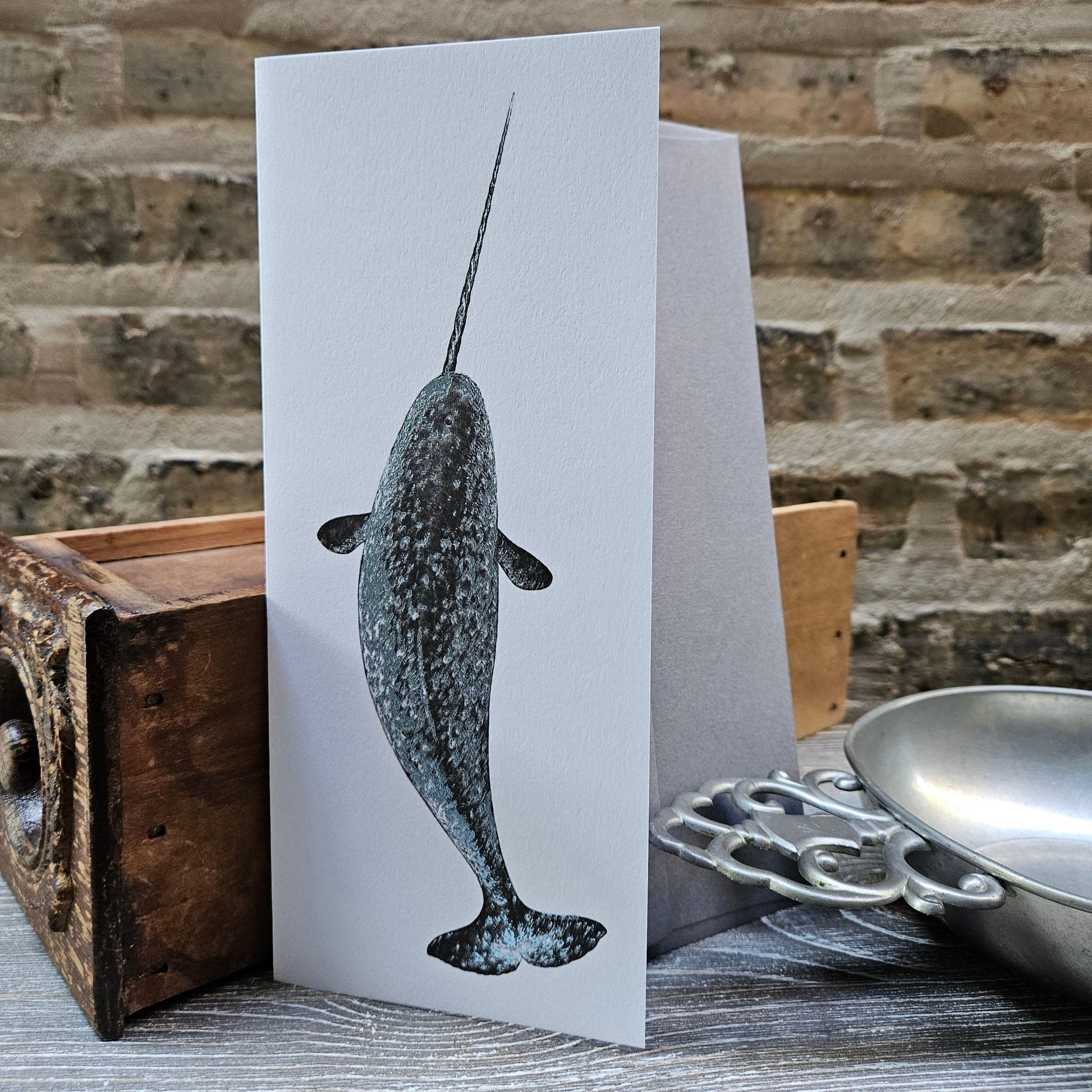 white, tall, rectangular card with blue-grey foil narwhal illustration on front. Background is old brick and card sits along antique props.