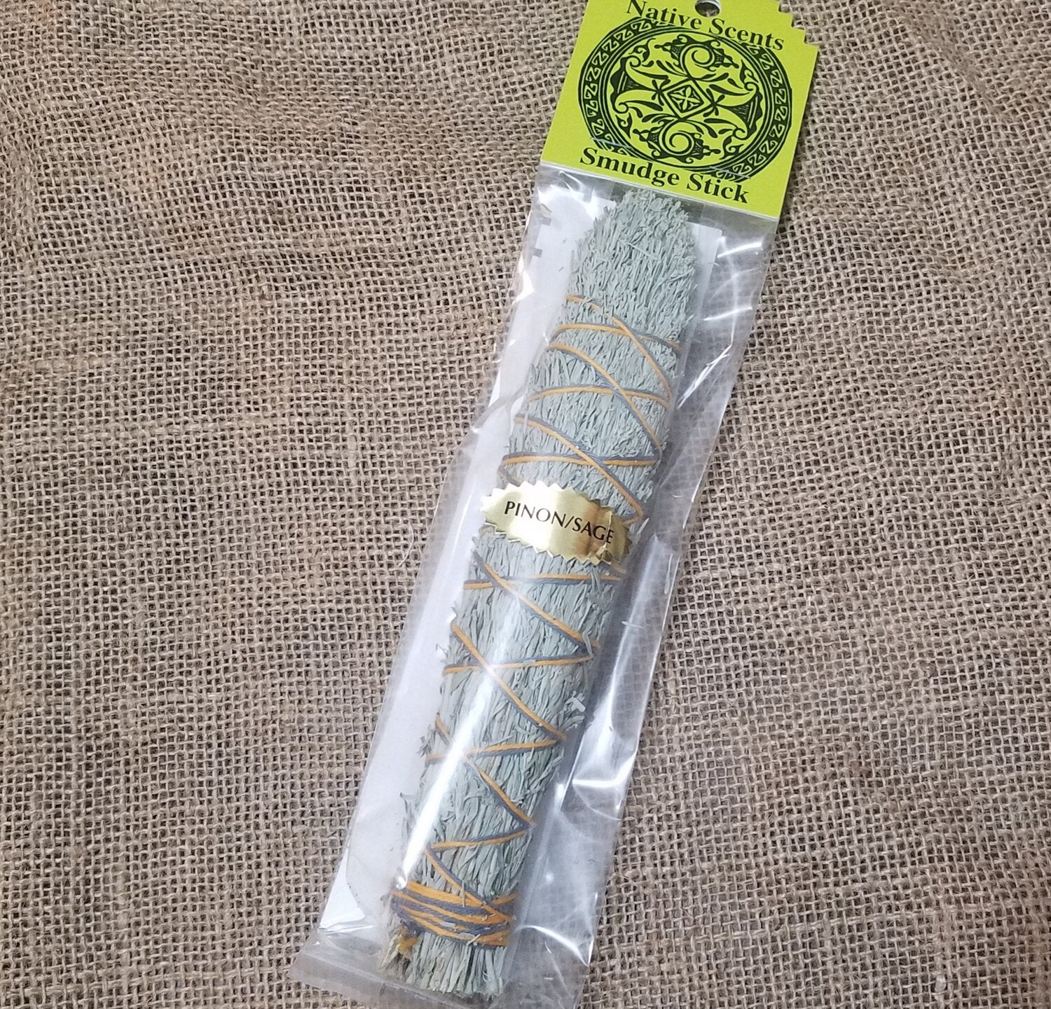 Large smudge stick wrapped in criss-cross string. Celophane packaging with lime green cardboard label and metallic gold sticker that reads "pinion/sage".  Background is burlap.