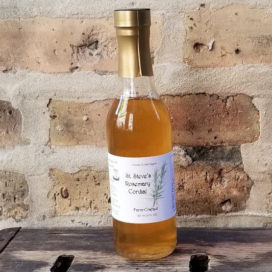 a tall thin clear glass bottle with amber liquid sits on a wooden platform in front of a brick background. bottle label is white with an illustration of a rosemary plant. bottle is sealed in a dull gold wrapper at the neck.